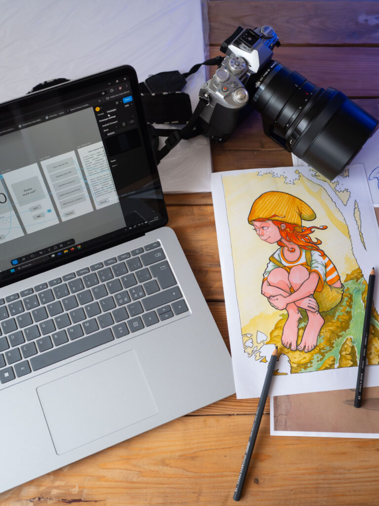 Photograph of a table with a laptop computer, drawings, pencils and a camera.